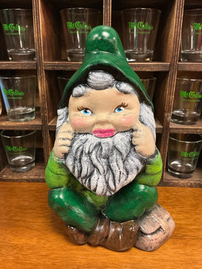 Decorative gnome figure with green hat and gray clothing, placed on a wooden surface with shot glasses in the background.