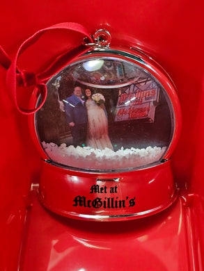 A red snow globe ornament with a photo of a couple and the text 'Met at McGillins'. It is adorned with a red ribbon.