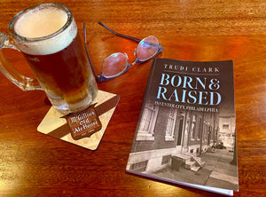 A book titled 'Born & Raised' by Trudi Clark, placed on a table with a glass of beer and sunglasses, set against a backdrop of a Philadelphia cityscape.